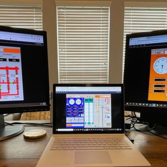 A desk with two large monitors displaying system control interfaces and a laptop in the center also showing a control interface. Three windows with closed blinds are in the background.