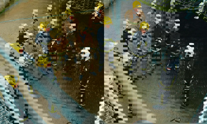 Um grupo de bombeiros vestidos com capacetes amarelos e equipamento de combate a incêndios está em formação, ouvindo outro bombeiro de capacete branco a dar instruções.