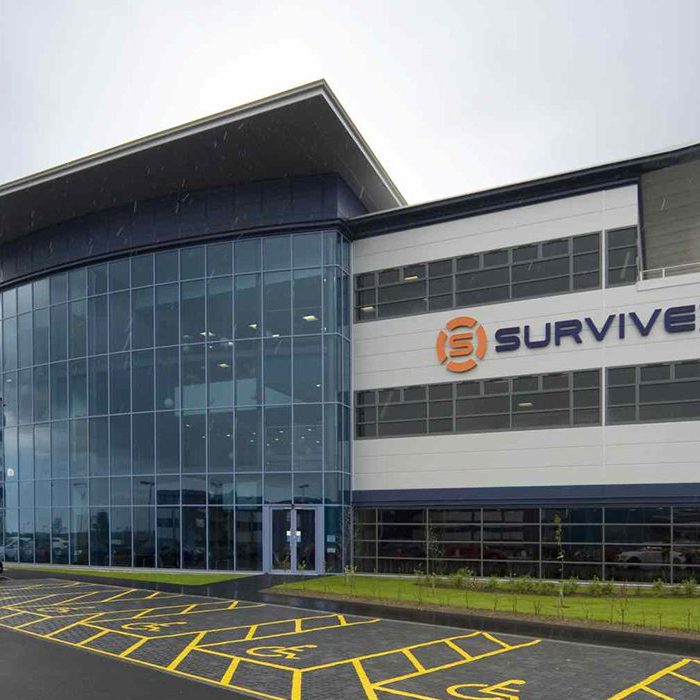 Modern office building with glass facade and "Survivex" sign. Several cars are parked in the lot, with marked yellow lines on the pavement. Overcast sky in the background.