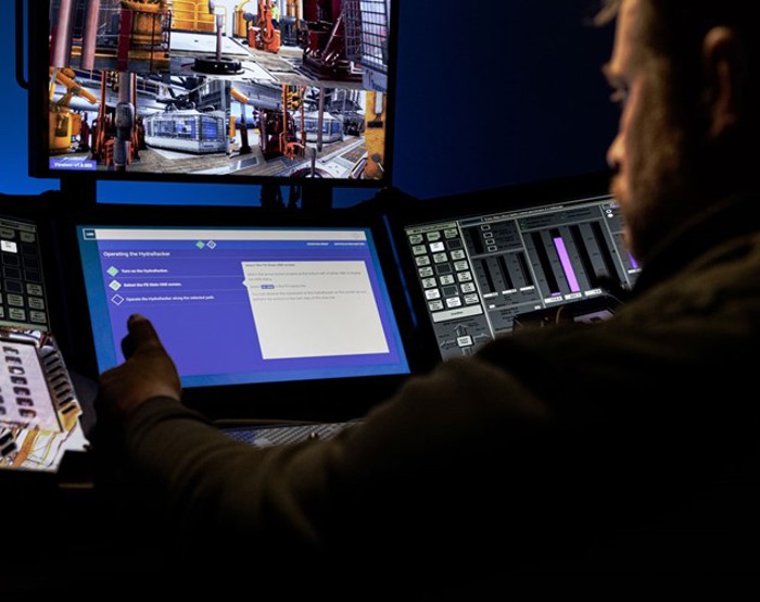 Uma pessoa opera um equipamento utilizando vários ecrãs, incluindo um que mostra imagens de máquinas e outro que apresenta dados e controlos digitais.
