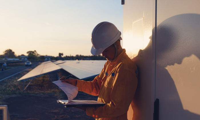 Un ouvrier en uniforme jaune et casque de chantier lit des documents à l'extérieur, au coucher du soleil, adossé à un mur à côté de panneaux solaires.