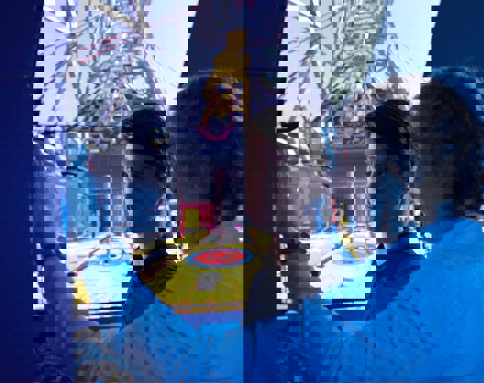 Dois homens numa sala de controlo operam um simulador com um grande ecrã que mostra o interior de um ambiente industrial. Um deles está a apontar enquanto o outro observa atentamente.