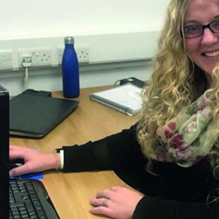 A person with long curly hair and glasses is sitting at a desk, smiling while using a computer. There is a blue water bottle, a notebook, and other office items on the desk.