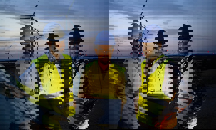 Trois travailleurs en tenue de sécurité et casqués suivent une formation alors qu'ils examinent un ordinateur portable près d'éoliennes au crépuscule.